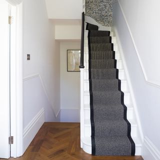 Brown wooden floor leading to white stair case covered with black stair case runner