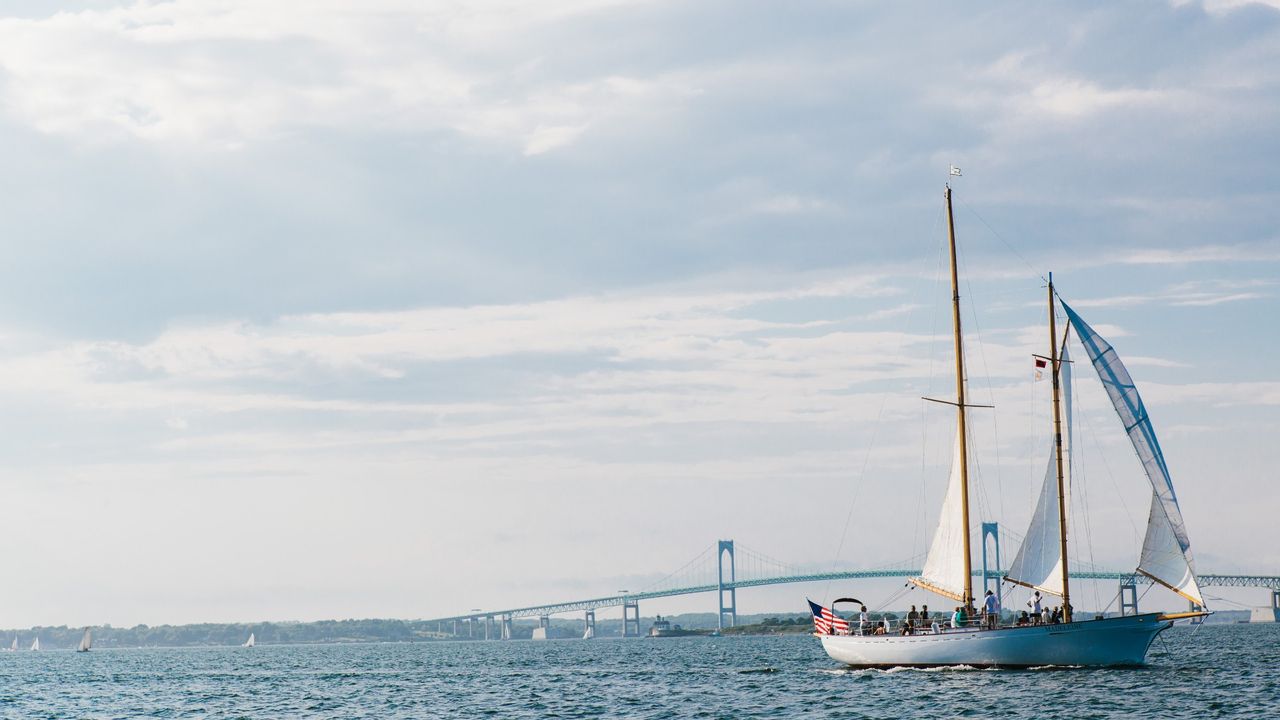 A white sailboat sails through Rhode Island&#039;s Newport Harbor on a clear day with the Newport Bridge in the background