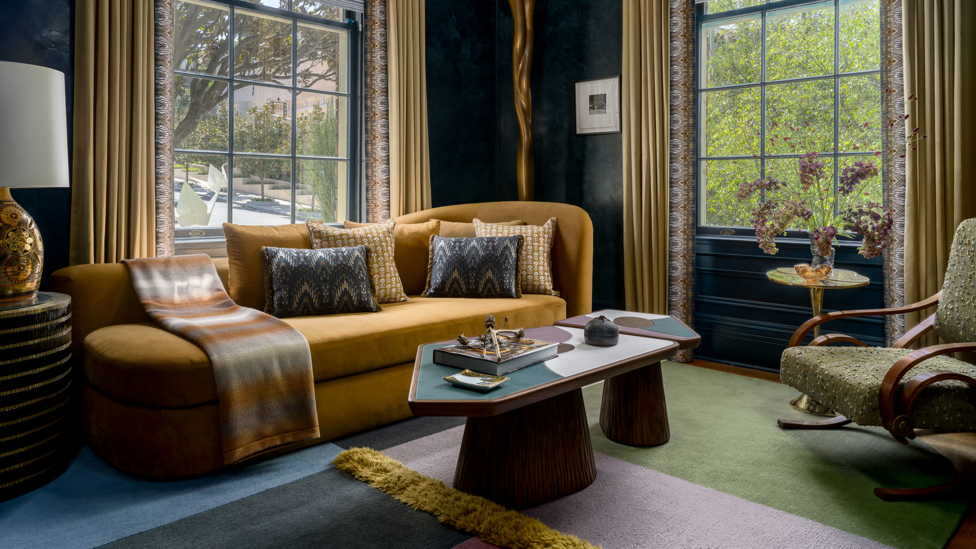 An eclectic living room with navy textured walls, a mustard velvet sofa, and two geometric coffee tables. Large windows with gold drapes frame a bright view, complementing a colorful paneled rug.