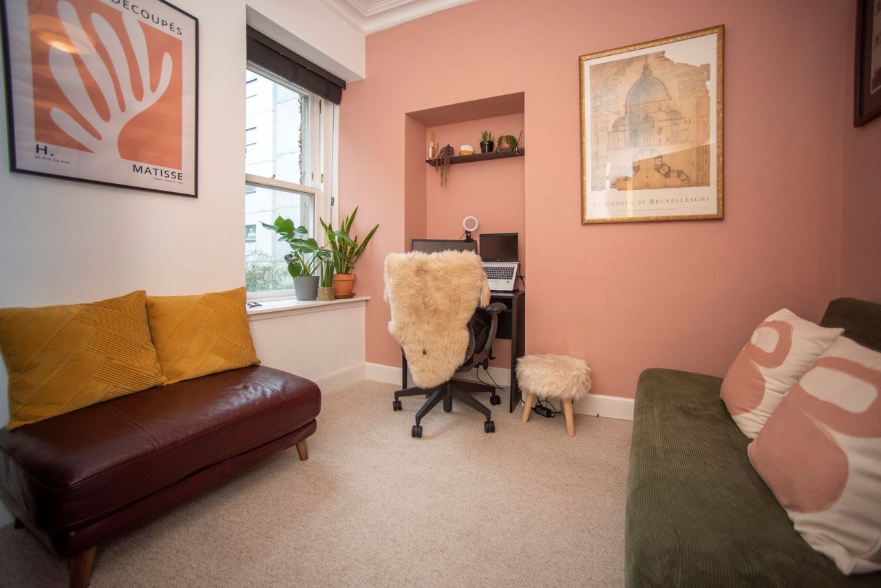 The office has pink walls and the computer is sat next to a large window with more plants on the window sill and includes leather sofas