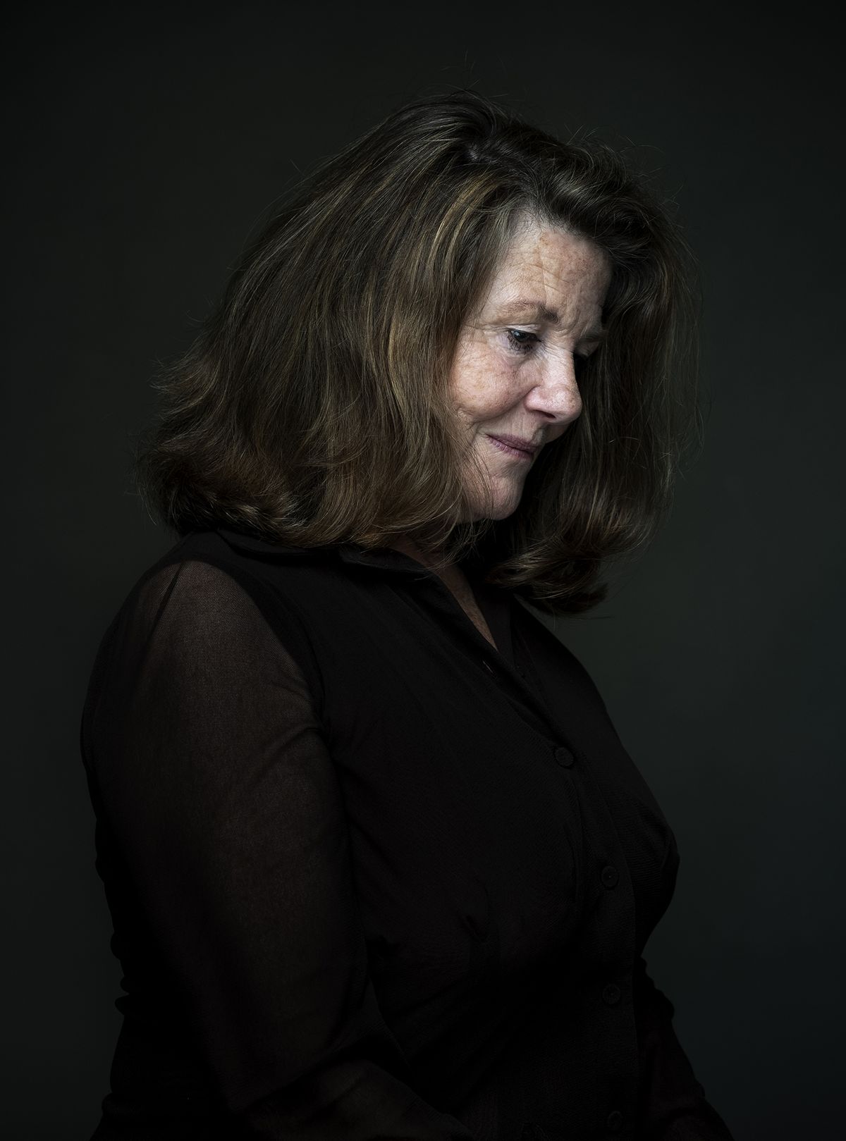 A woman with wavy, shoulder-length hair wearing a black, sheer blouse, against a dark background, striking a contemplative pose