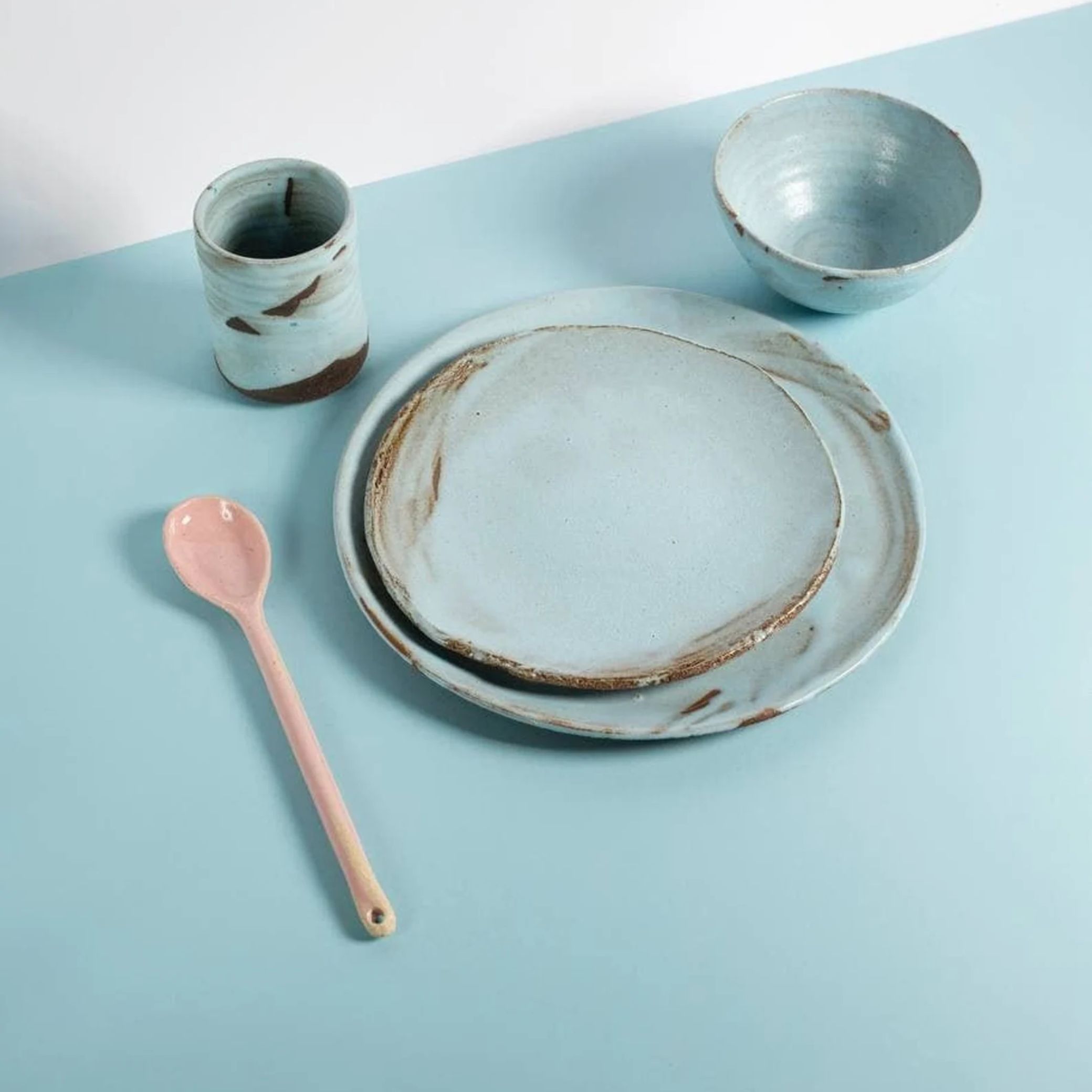 A Rustic Dinnerware Set in pale blue ceramic sits atop a pale blue surface next to a pale brown, ceramic spoon.