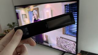 En person holder en fjernbetjening til Samsung The Frame TV 2021 med selve TV'et i baggrunden.