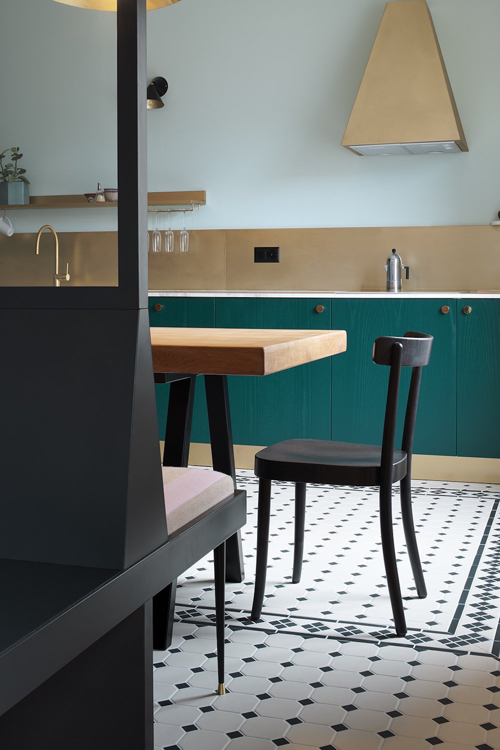 A view of a kitchen with pale blue walls, gold backsplash, marble work tops, teal cupboards and a black bench seating area.