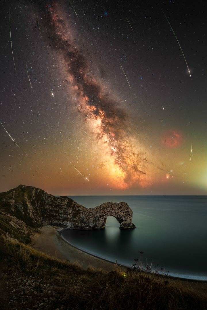 A night sky image shows the glowing streak of the Milky Way galaxy over a stone arch on a coastline, with streaks of white meteors in the sky.