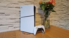 The PS5 Slim and DualSense controller on a wooden table in front of a white brick background and next to a vase of flowers