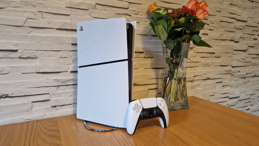 The PS5 Slim and DualSense controller on a wooden table in front of a white brick background and next to a vase of flowers