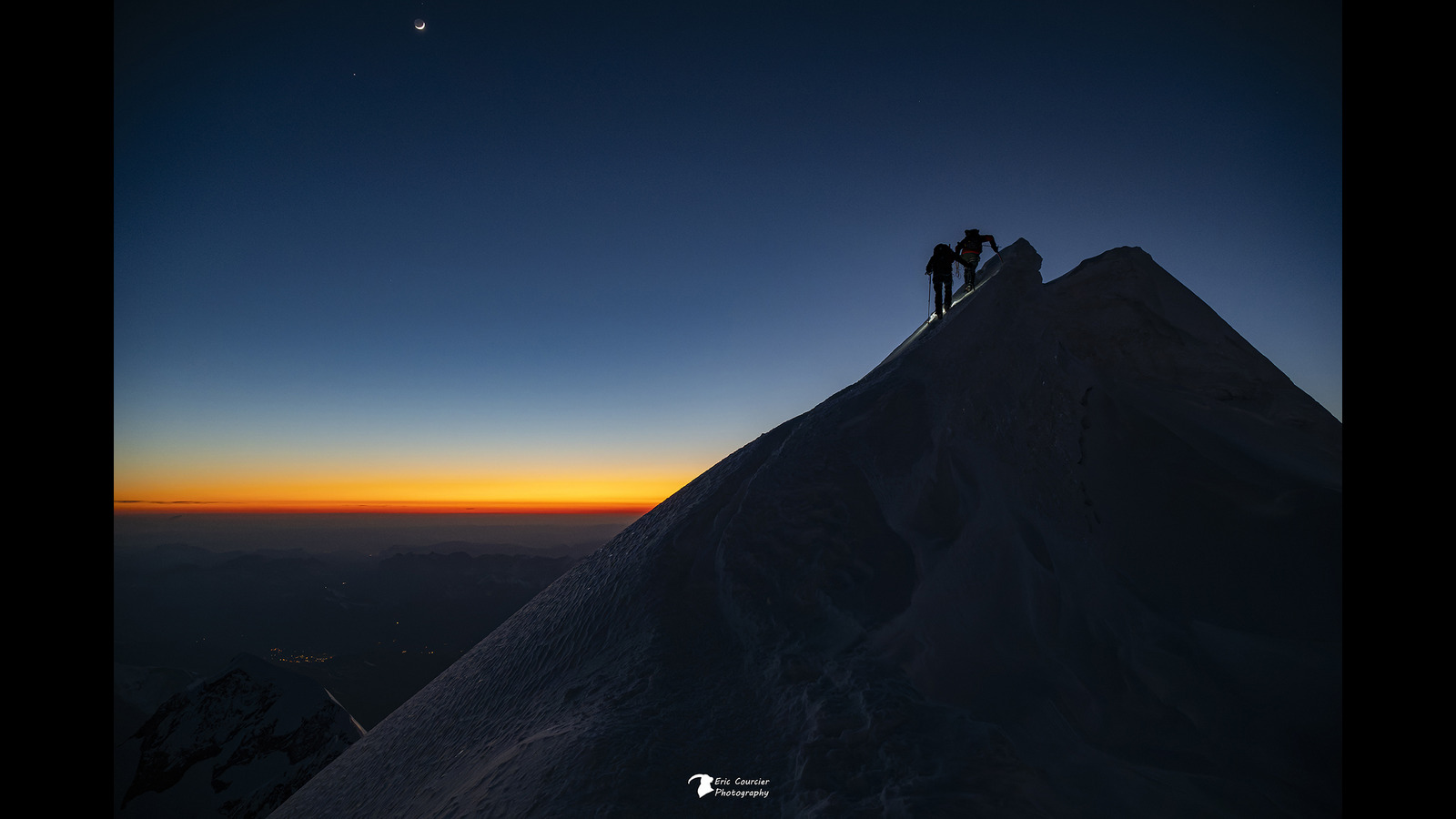 Two people hike on a mountain at dusk with a crescent moon overhead