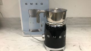 smeg milk frother in black in front of the box on a marble countertop