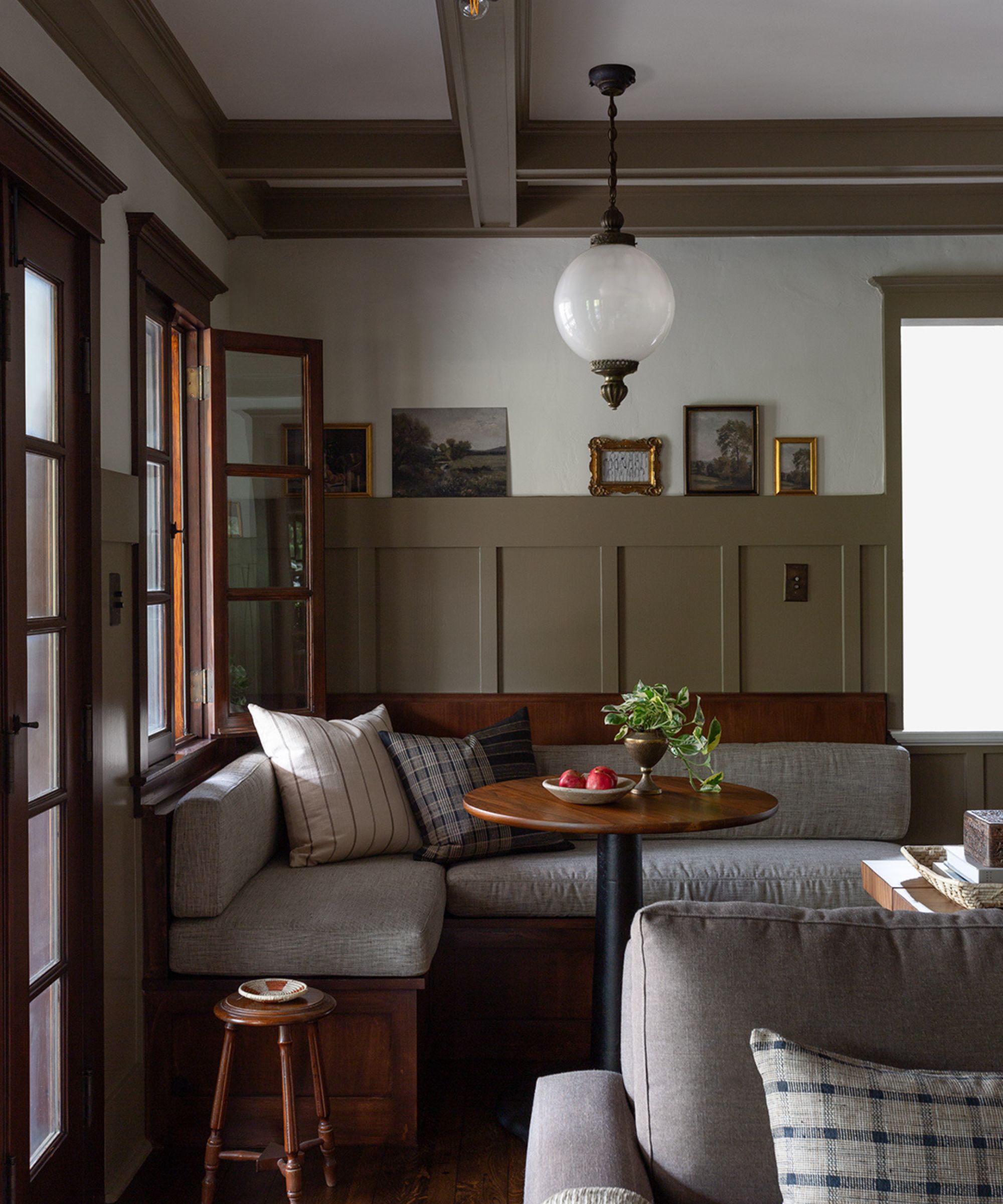 Breakfast nook with wall panelling and beams painted in Mouse's Back by Farrow &amp;amp; Ball, a built-in wooden banquette with a grey cushion, a round wooden table, and a glass antique pendant light.