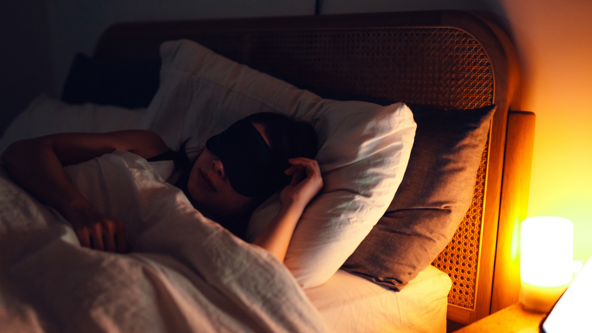 Woman sleeping in a cozy bedroom with warm lighting with two pillows under her head, an eye mask and a comforter