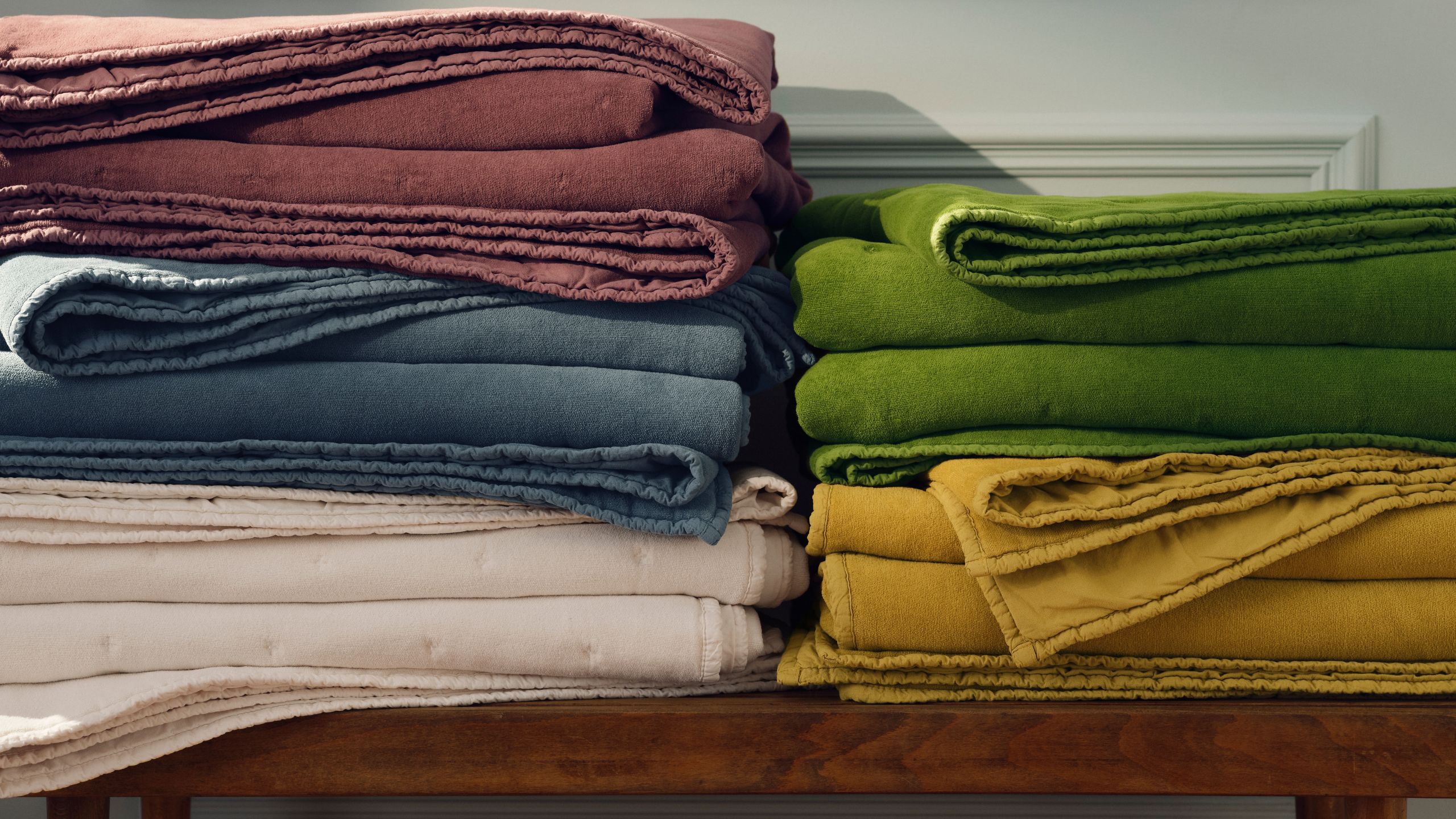 Five Brooklinen Washed Velvet Quilts in every color folded up on a wooden bench.