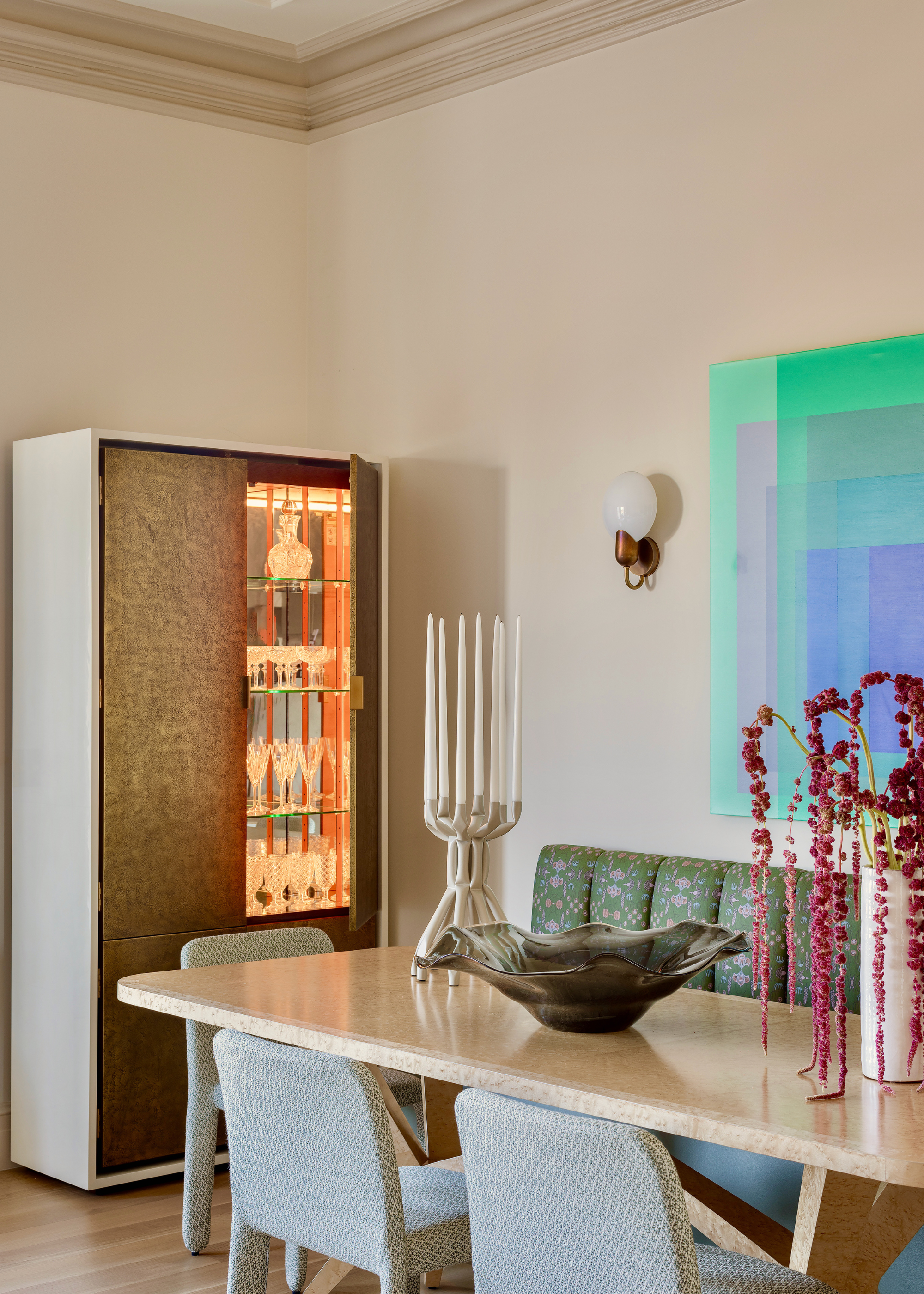 A bar cabinet next to a dining table with banquette seating has one door open revealing lit shelving filled with glassware