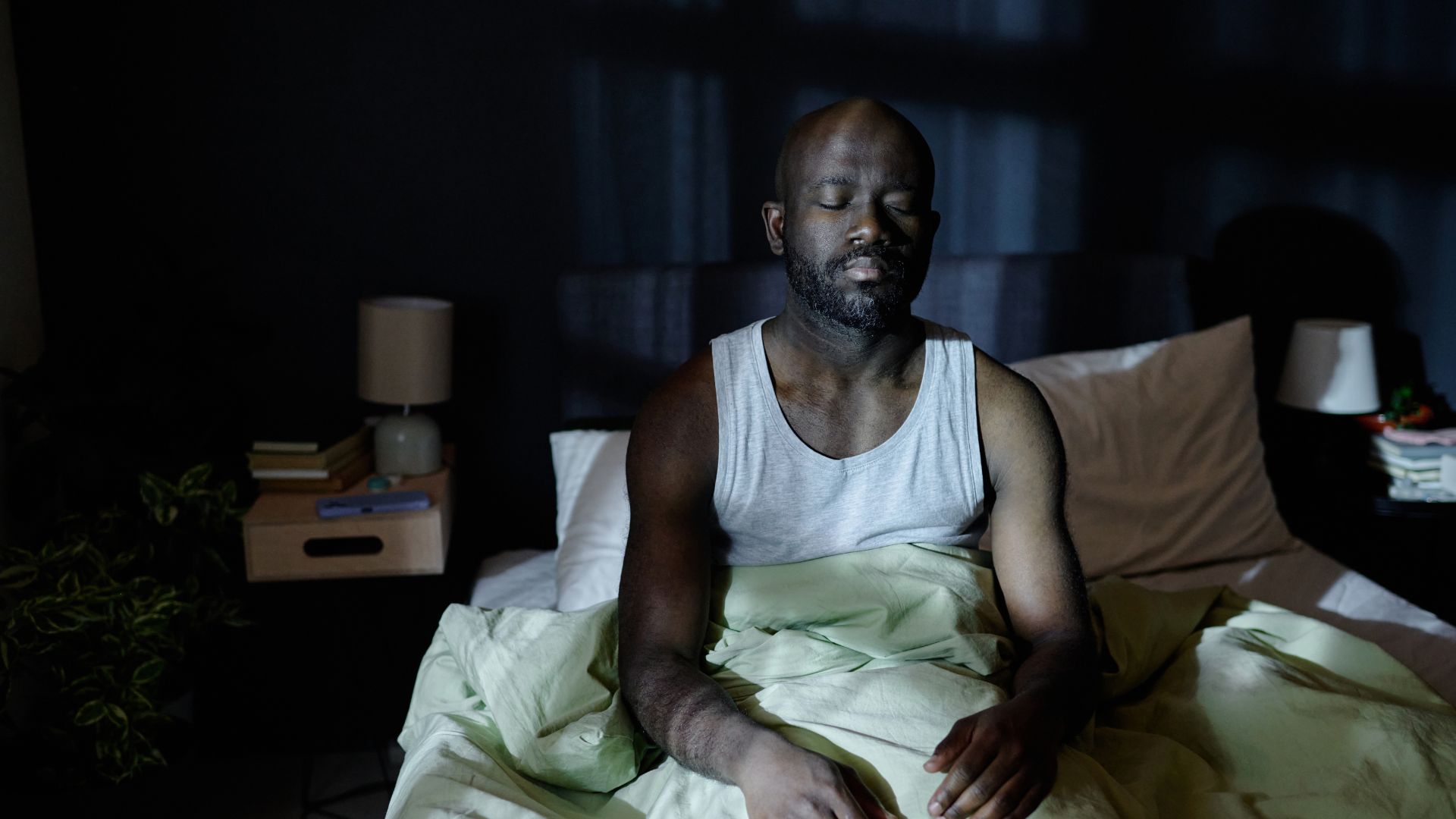 A man sits up in bed in a darkened room with his eyes closed.