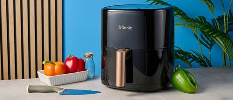 a large black 5.3 quart capacity air fryer with black touchscreen and rose gold embellishments sits upon a brown table with a blue background with red bell peppers and a glass jar next to it