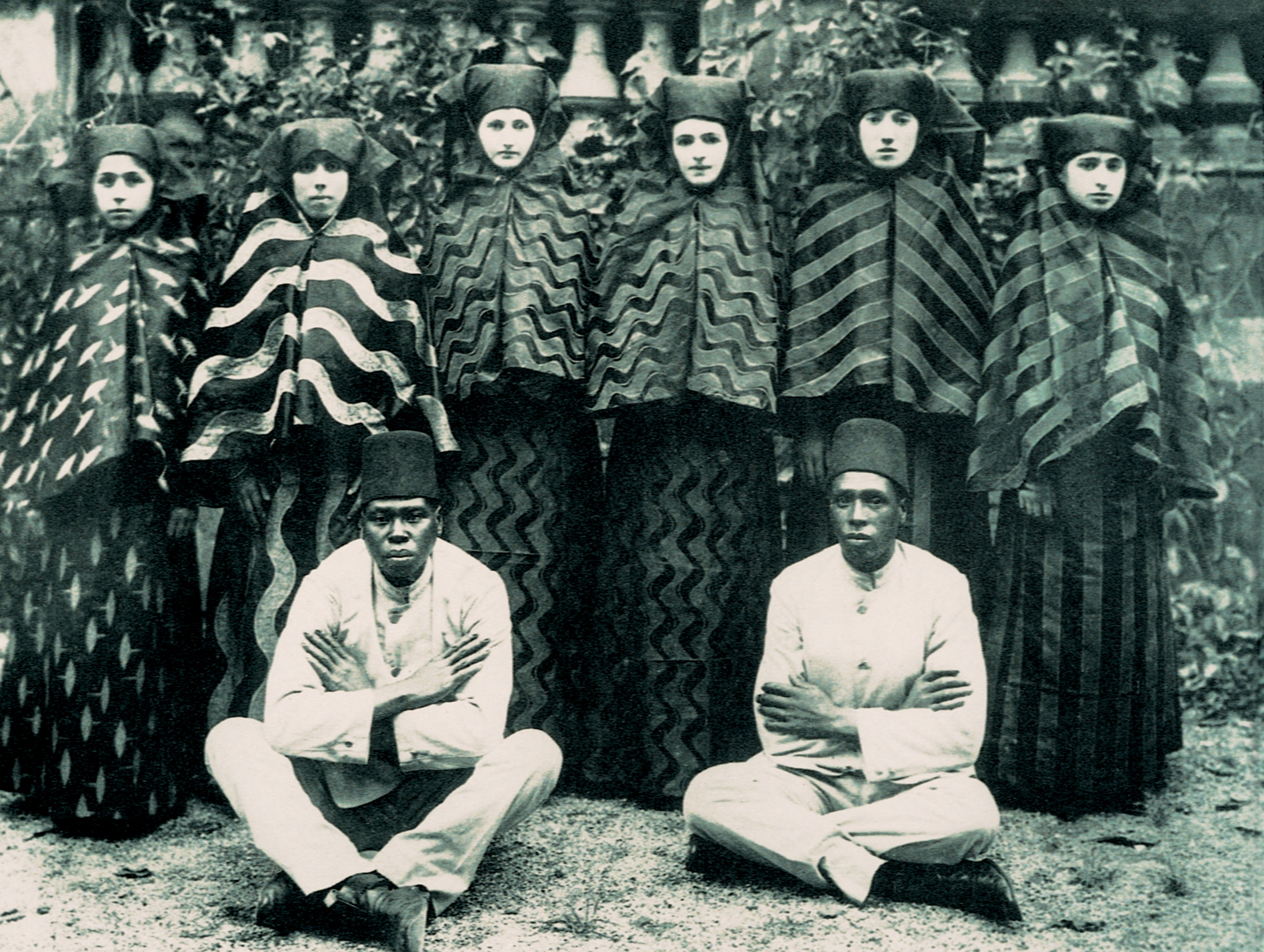 A black and white photograph shows six women standing wearing full coverings of black and white while in the foreground two men wearing tall flat black hats and white clothing sit cross legged.