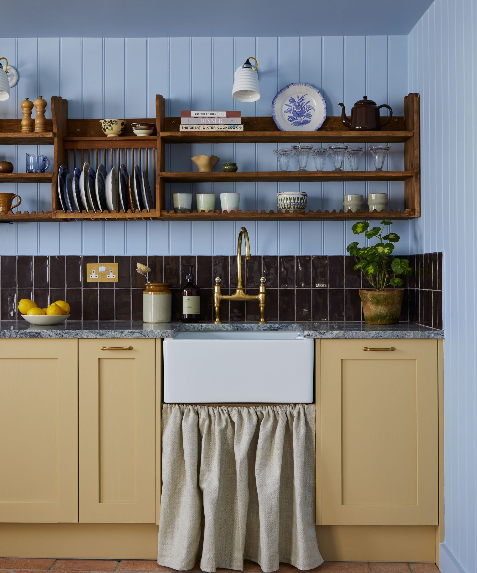 A cozy cottage kitchen with powder blue color-drenched panelled walls and butter yellow kitchen cabinets. Open wood shelving and dark burgundy backsplash tiles.