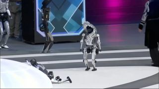 A robot wearing a hat watching another robot fall over at the World Humanoid Robot Games