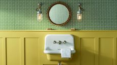 A yellow sink on a yellow panelled half wall, green patterned wallpaper on the wall above. A small round mirror flanked by two small pendant lights. A small folded white face cloth over the side of the sink