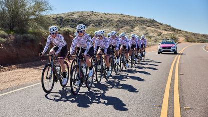 A peloton of the DNA Pro Cycling women's team riding on a road.