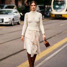 woman wears white sweater, asymmetrical handkerchief hemline dress with tall boots and a matching bag