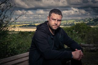 Emmerdale favourite Aaron Dingle sitting on a bench with Yorkshire fields behind him.