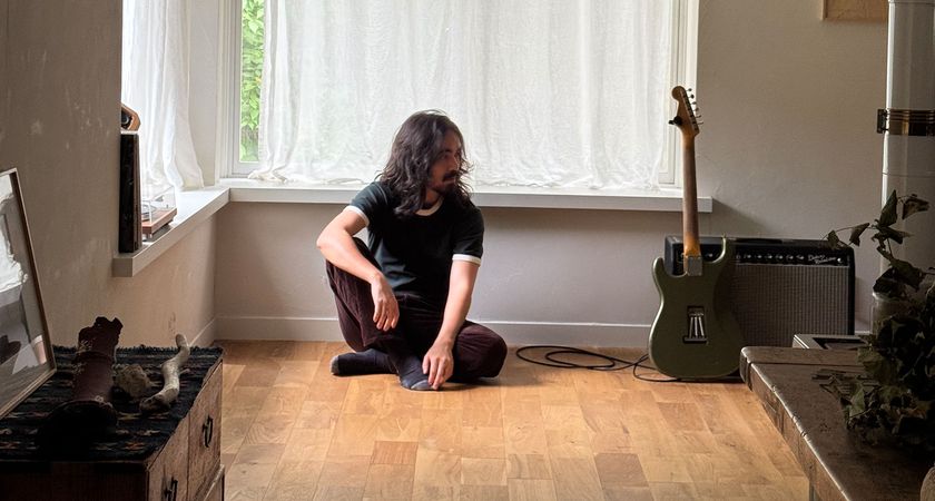 Mateus Asato sits on a wooden floor, while a Strat leans against an amp.