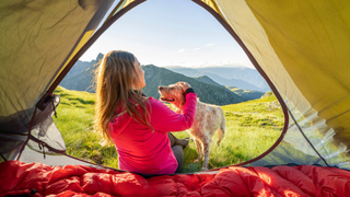Woman and dog sitting outside of a tent with a mountain view behind them