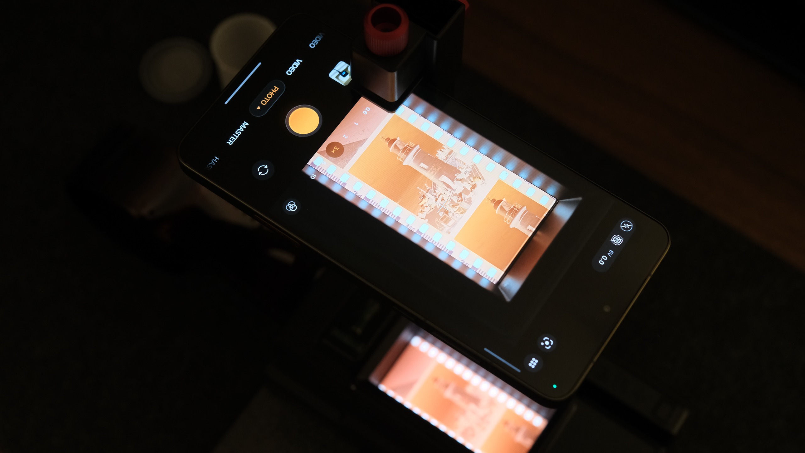 A top-down view of a smartphone mounted on a scanning stand, with its camera app open and aligned to photograph a backlit strip of 35mm film displaying a negative of a lighthouse.