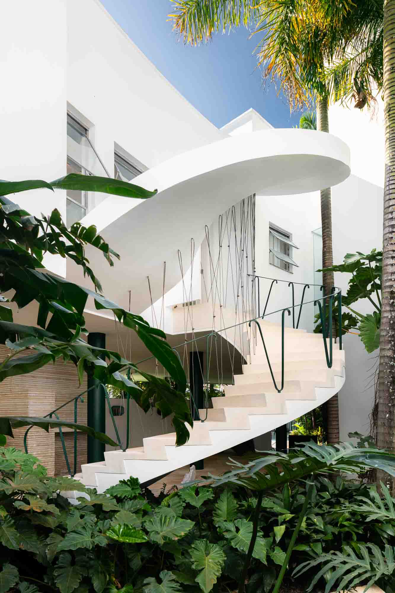 The exterior of a modernist, white-washed tropical building is immersed in green vegetation and decorated with a twisting staircase with iron details on a sunny day.