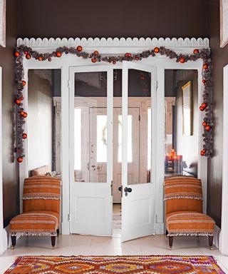 Green foliage garland with red orange Christmas decorations in an entrance hall with porch, double doors and front door.