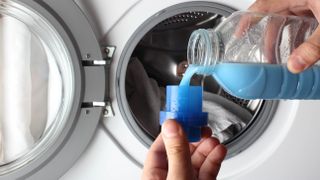 A person measuring out fabric softener in front of a loaded washing machine