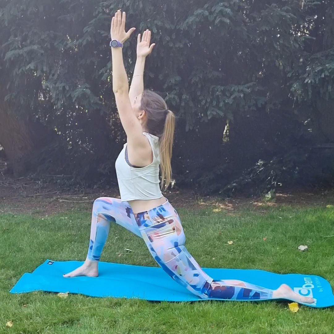 Fitness editor Ruth Gaukrodger performs a low lunge to half split. She is in sportsgear and exercising outside on a yoga mat. In this picture, she is performing a low lunge. Her right knee is bent at a right angle in front of her, with her foot planted on the ground, while her left leg lies behind her. Her arms are held up towards the sky.