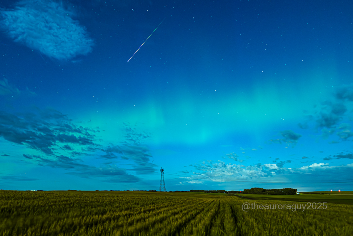 Meteors are visible streaking through a dark blue sky wreathed in a green aurora above a rural landscape.