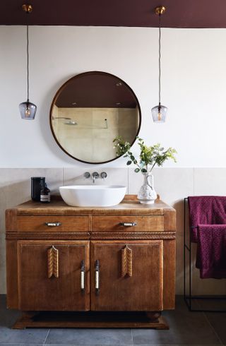Bathroom makeover: shot of vanity unit in bathroom. Deep wood vanity unit with oval sink plumbed in and round mirror above, framed by two pendant lights suspended from a purple-painted ceiling