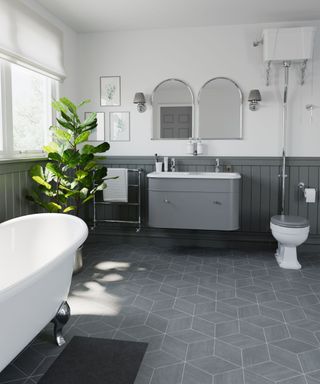 A bathroom with low level gray wall paneling, gray toilet lid, sink unit, two arched mirrors on the wall, a tall fiddle leaf fig plant, a freestanding bath with silver ornate feet and geometric gray floor tiling
