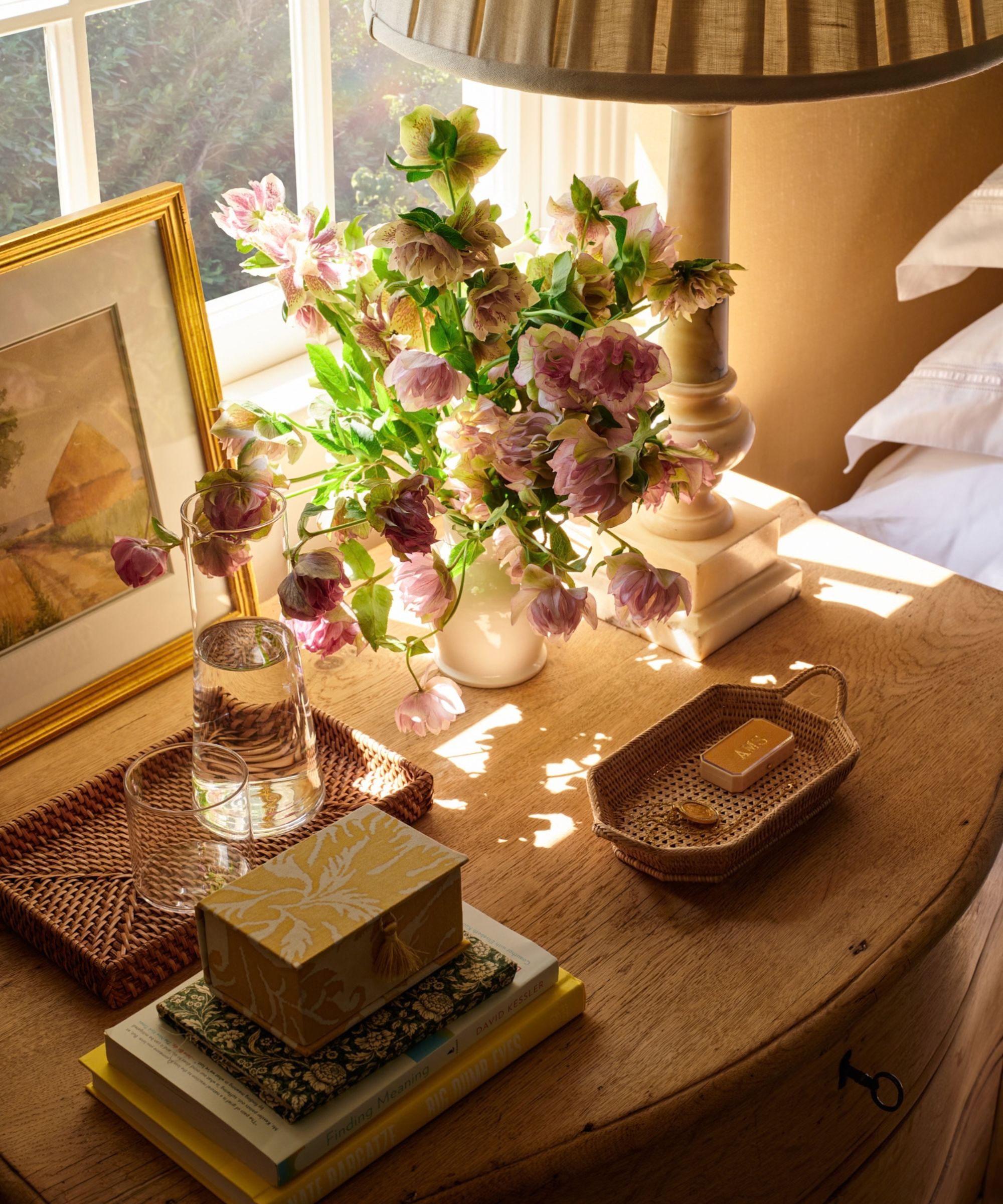 a bedside table vignette in front of a sunny window with wooden nightstand styled with a lamp, art, vase of flowers, wicker trays and trinkets