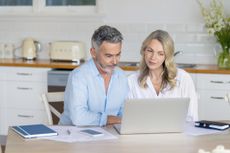 Mature couple doing paperwork and paying bills at home. They are using a laptop with the kitchen in the background.