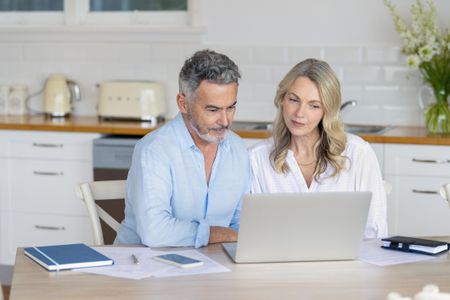 Mature couple doing paperwork and paying bills at home. They are using a laptop with the kitchen in the background.