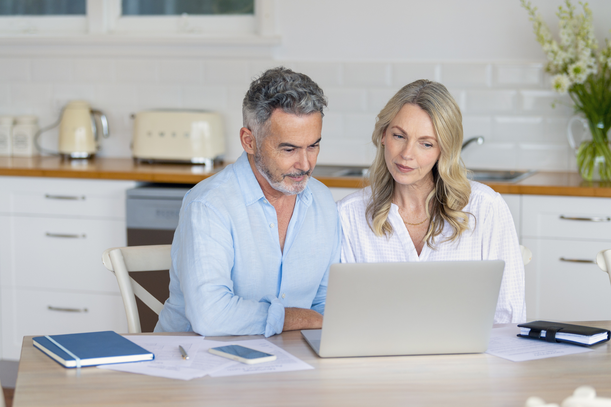 Mature couple doing paperwork and paying bills at home. They are using a laptop with the kitchen in the background.