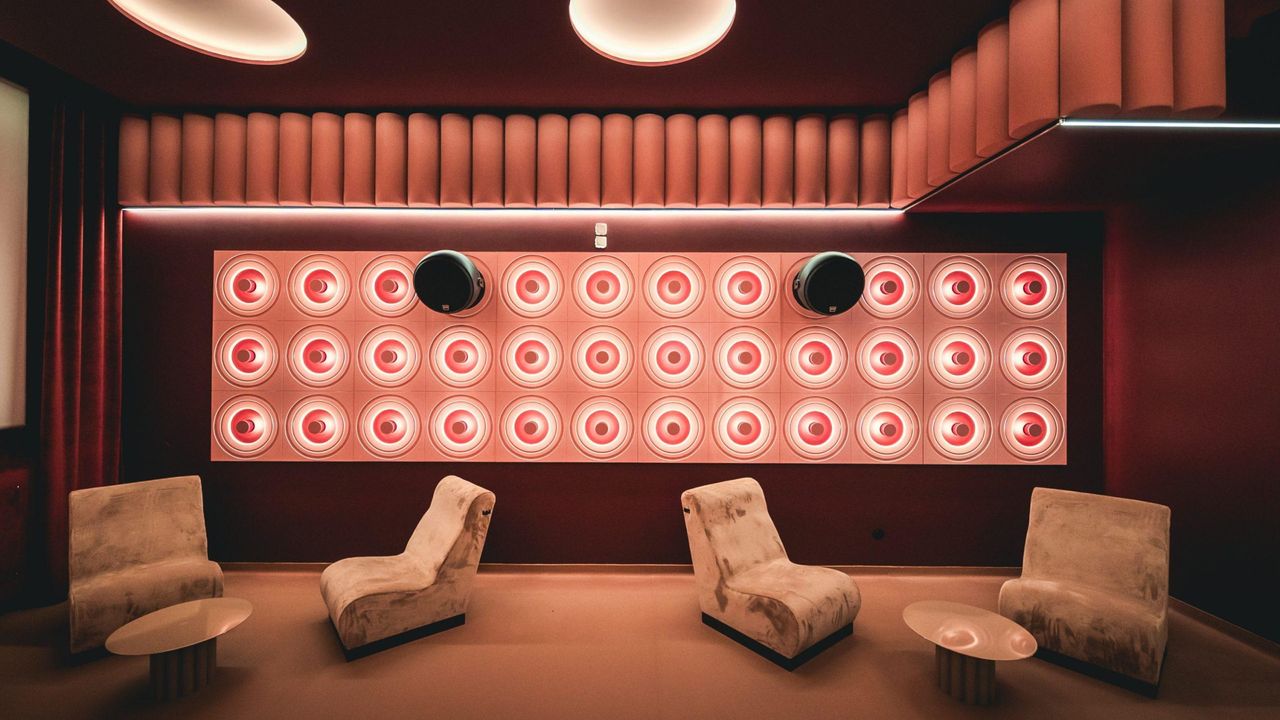 A retro-futuristic, boldly lit cinema room with high-tech sound panels in bright pink, velvety red curtains, cream velvet armchairs, round coffee tables in lacquered iron, and an immersive atmosphere.