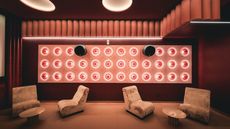 A retro-futuristic, boldly lit cinema room with high-tech sound panels in bright pink, velvety red curtains, cream velvet armchairs, round coffee tables in lacquered iron, and an immersive atmosphere.