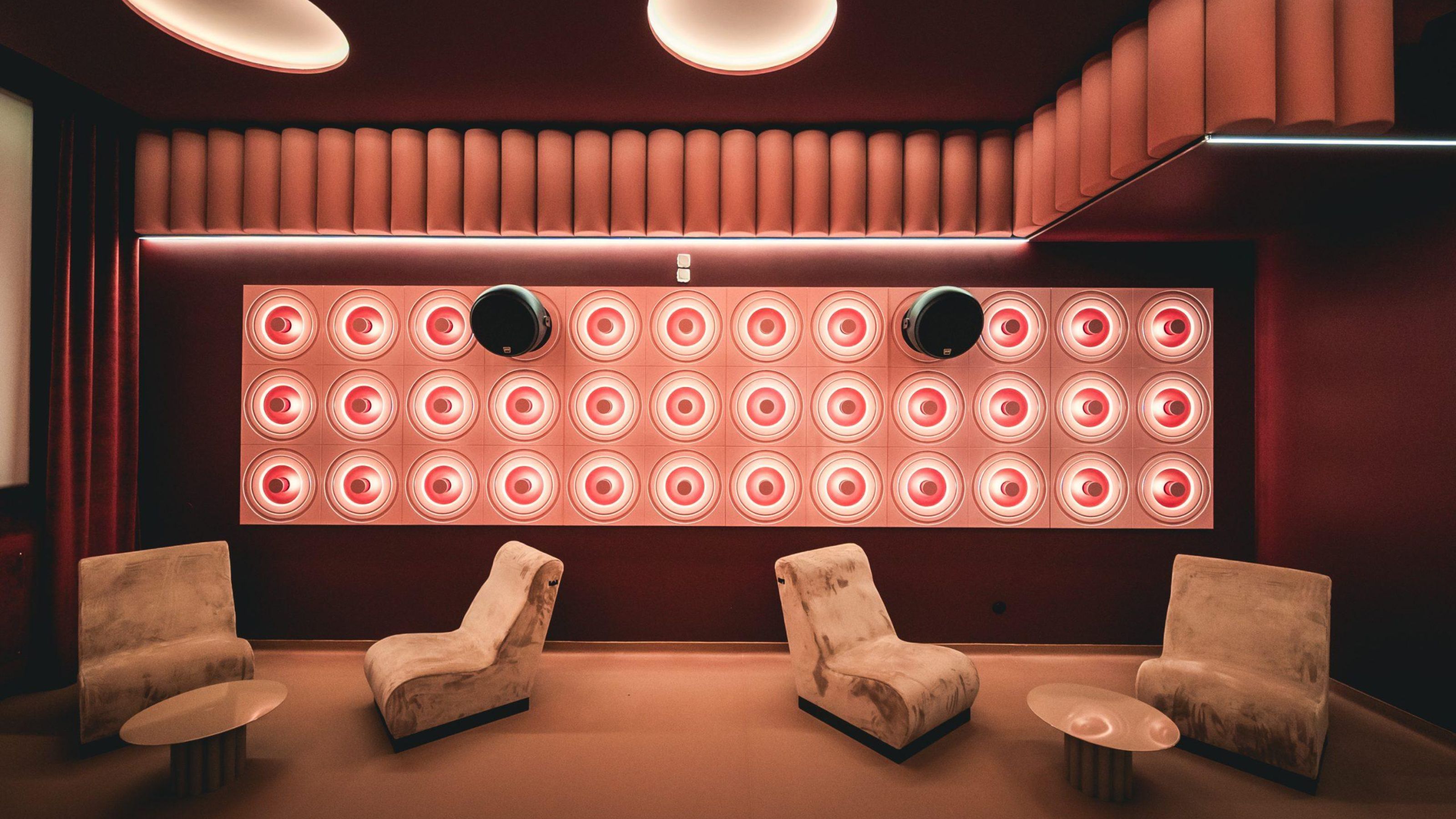 A retro-futuristic, boldly lit cinema room with high-tech sound panels in bright pink, velvety red curtains, cream velvet armchairs, round coffee tables in lacquered iron, and an immersive atmosphere.