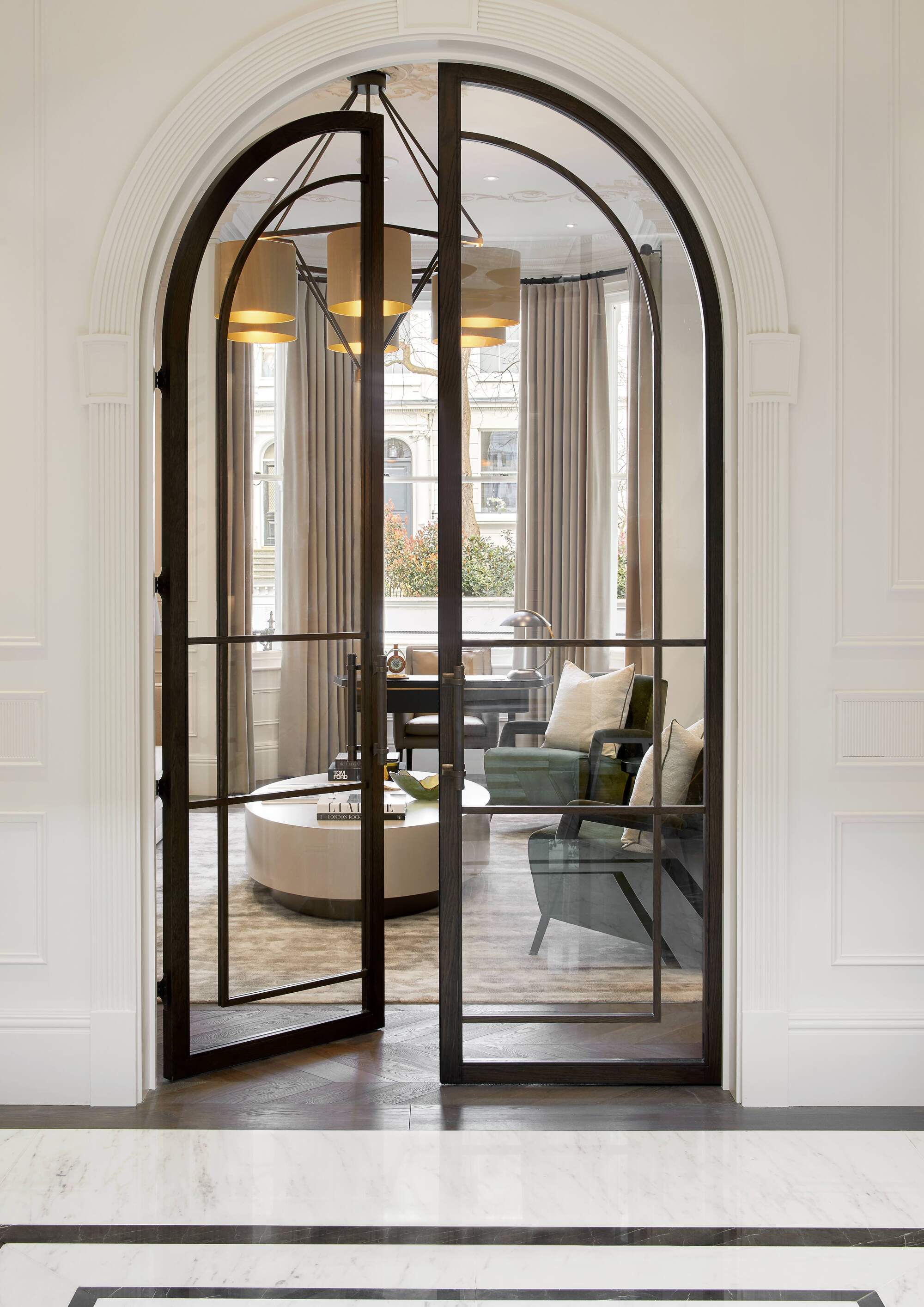 arched doorway with steel framed doors and wall molding leading through to a modern living room with a round coffee table, pendant light, and two blue armchairs with cushions