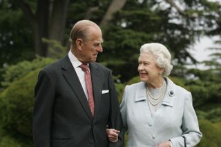 Queen Elizabeth wearing a blue coat and pearls holding arms with Prince Philip in front of trees outside