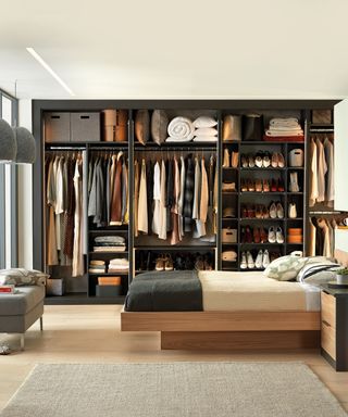 open closet with hanging and shelving sections, shoe racks, bedding on top shelf, modern wooden bed, ottoman, rug