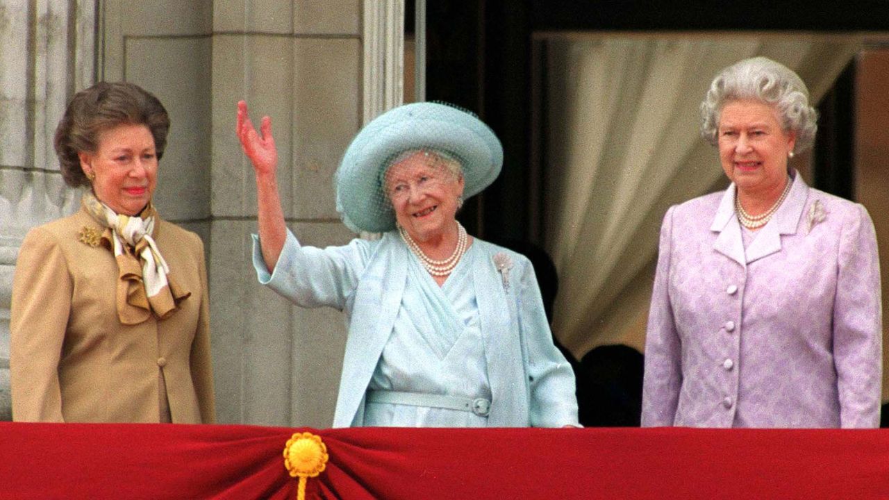 Princess Margaret, the Queen Mother wearing a blue hat and waving, and Queen Elizabeth wearing a pink suit standing on the balcony of Buckingham Palace