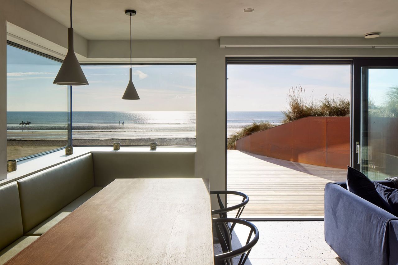 A view of the coast from the open plan living area with a dingin table and chairs in the foreground and bifolding doors open to the beach outside