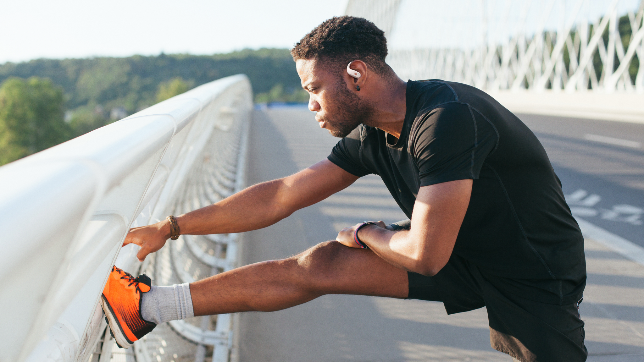 A young man with black hair, black t-shirt, black shorts and orange running shoes does a leg stretch on a white road bridge while wearing white JBL Endurance Zone headphones