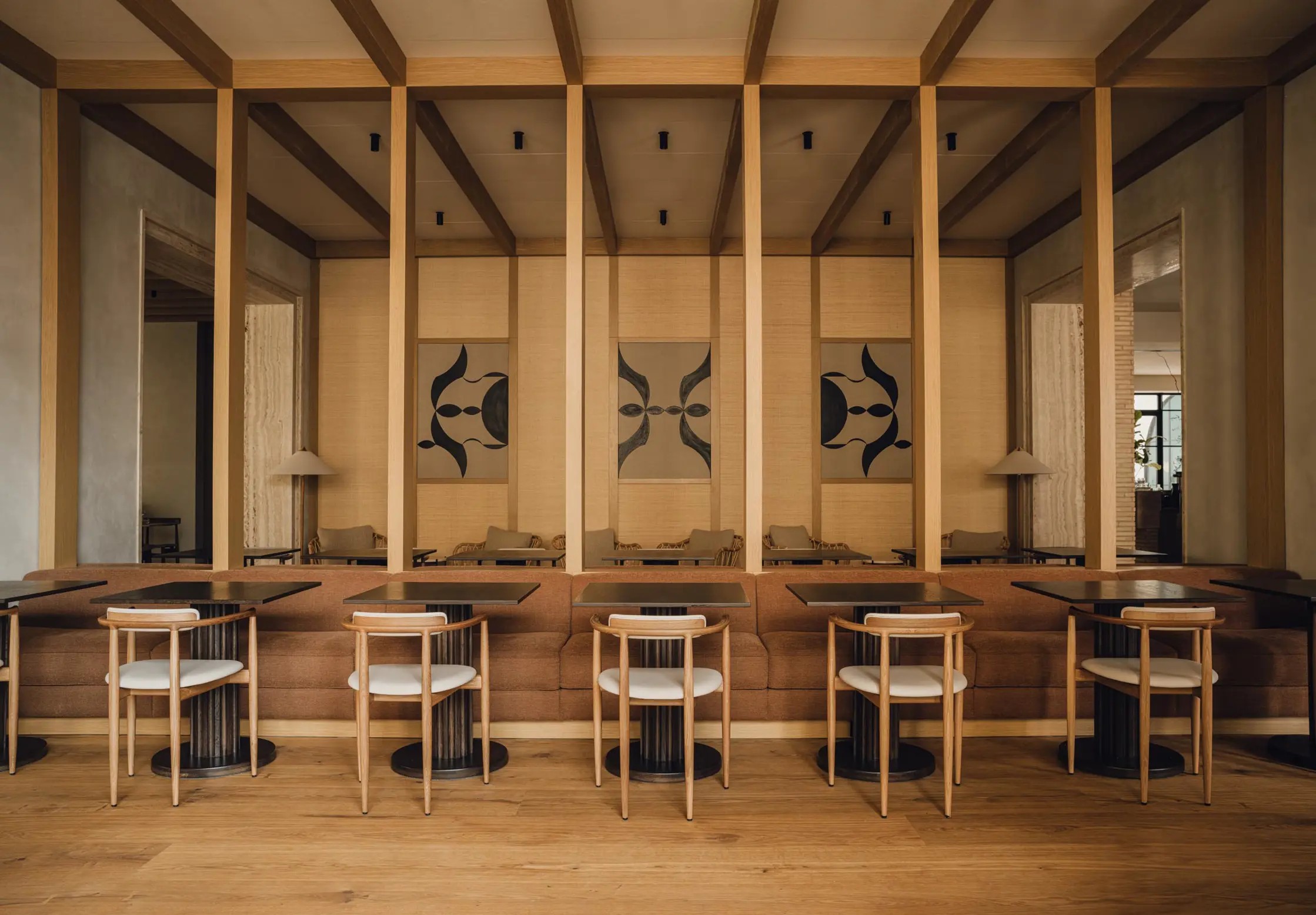 A wood-clad restaurant dining room with minimalist furnishings in wood, wrought iron, and brown velvet, and abstract brown and black paintings.
